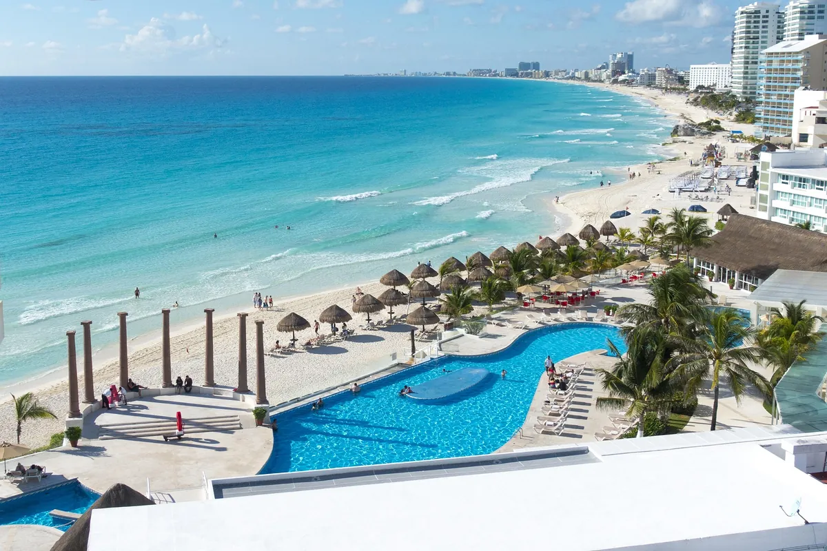 Rooftop resort pool overlooking the Cancun hotel zone coastline, with beach umbrellas, palm trees, and turquoise water below