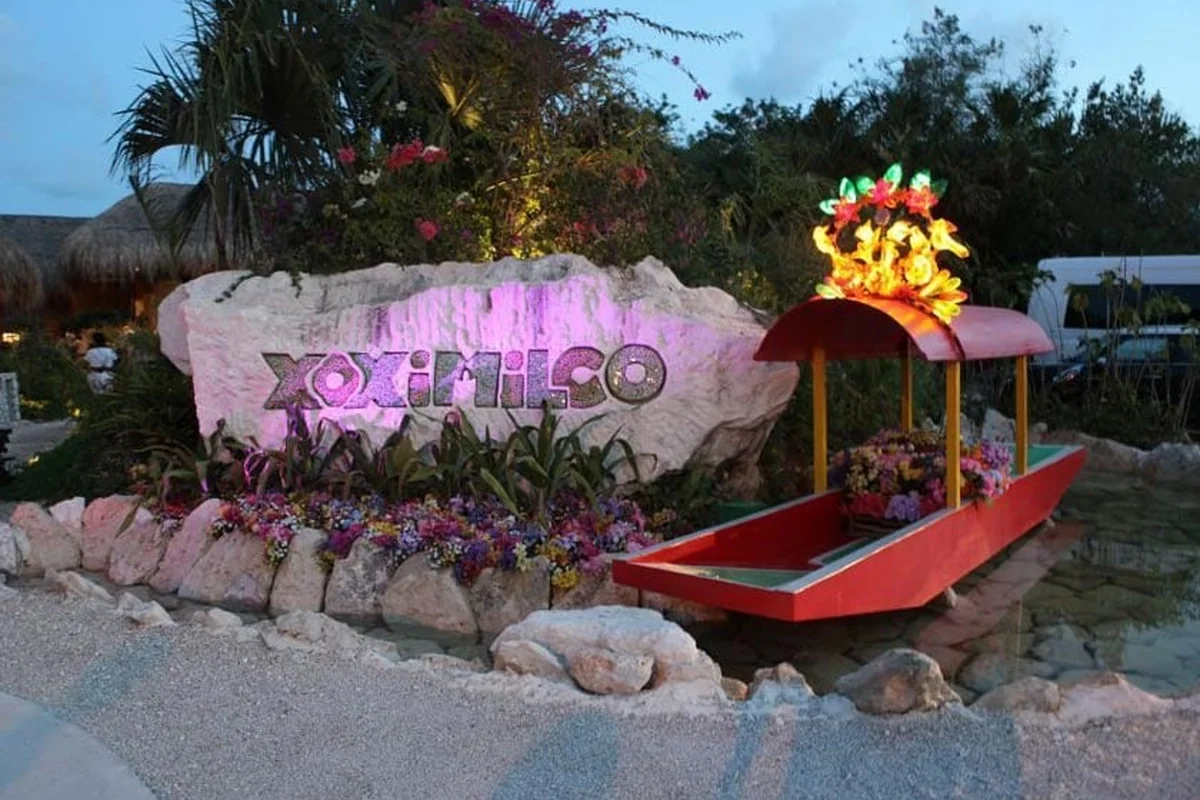 Illuminated Xoximilco entrance sign beside a flower-decorated trajinera boat at dusk