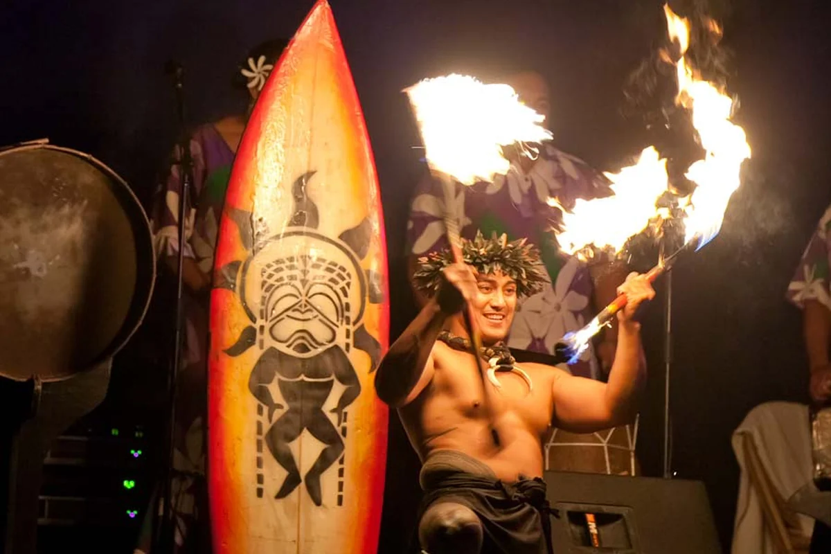 A fire dancer performing with flaming torches on stage at a nighttime Hawaiian cultural show, beside a hand-painted tiki surfboard