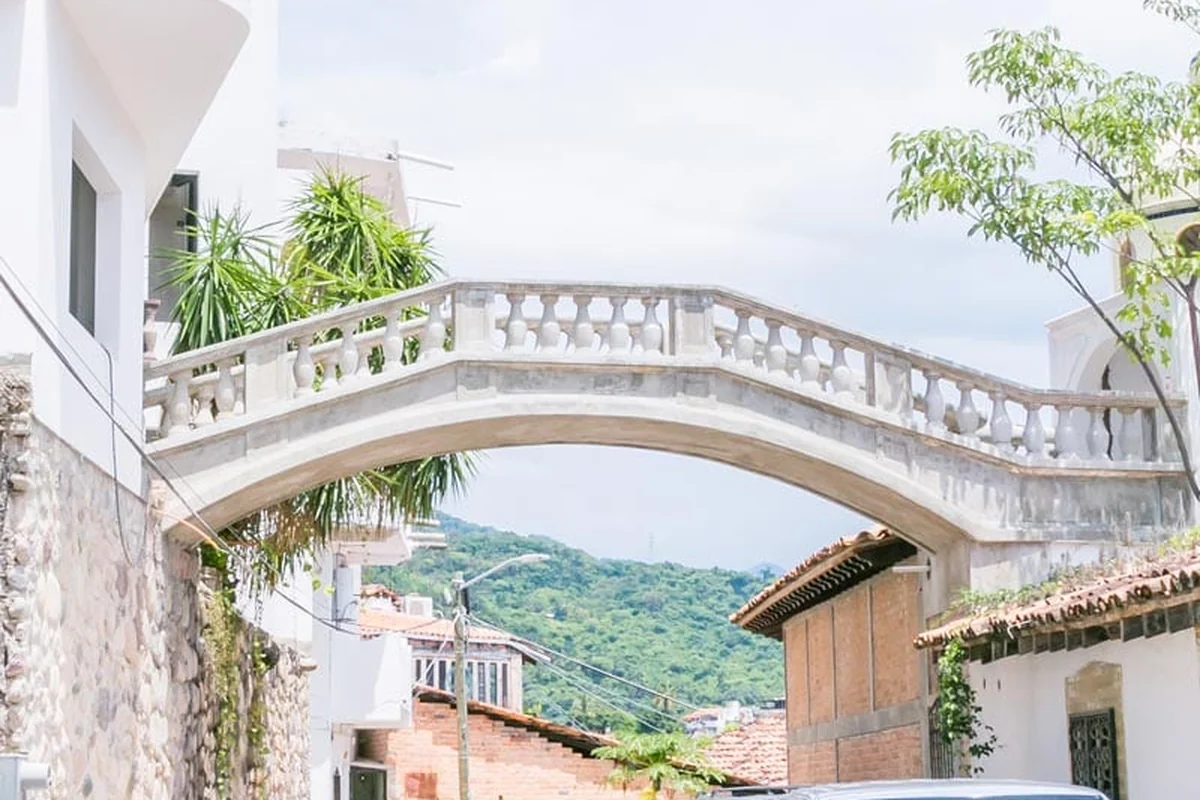 A carved stone footbridge arching over a street in Puerto Vallarta's old town, hillside and rooftops behind